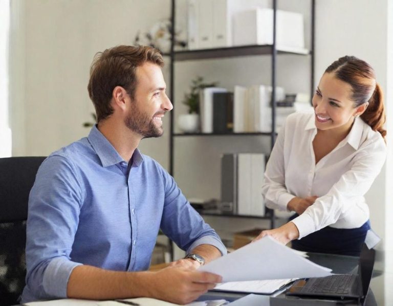 Zwei Kollegen im Büro Ein Mann am Schreibtisch, lächelt, während eine Frau ihm Dokumente reicht.